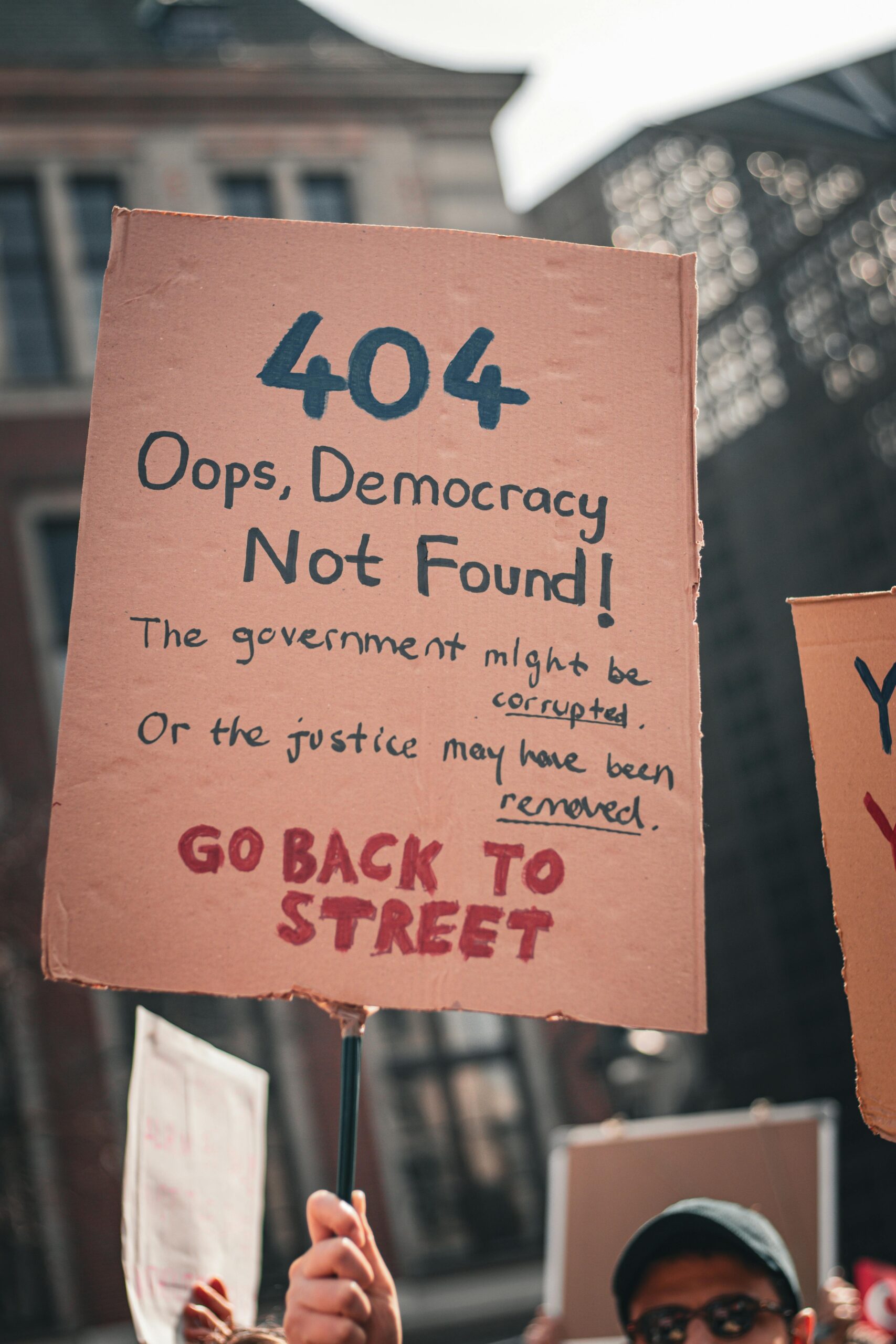 Protest sign reading '404 Oops, Democracy Not Found!' during a street demonstration.