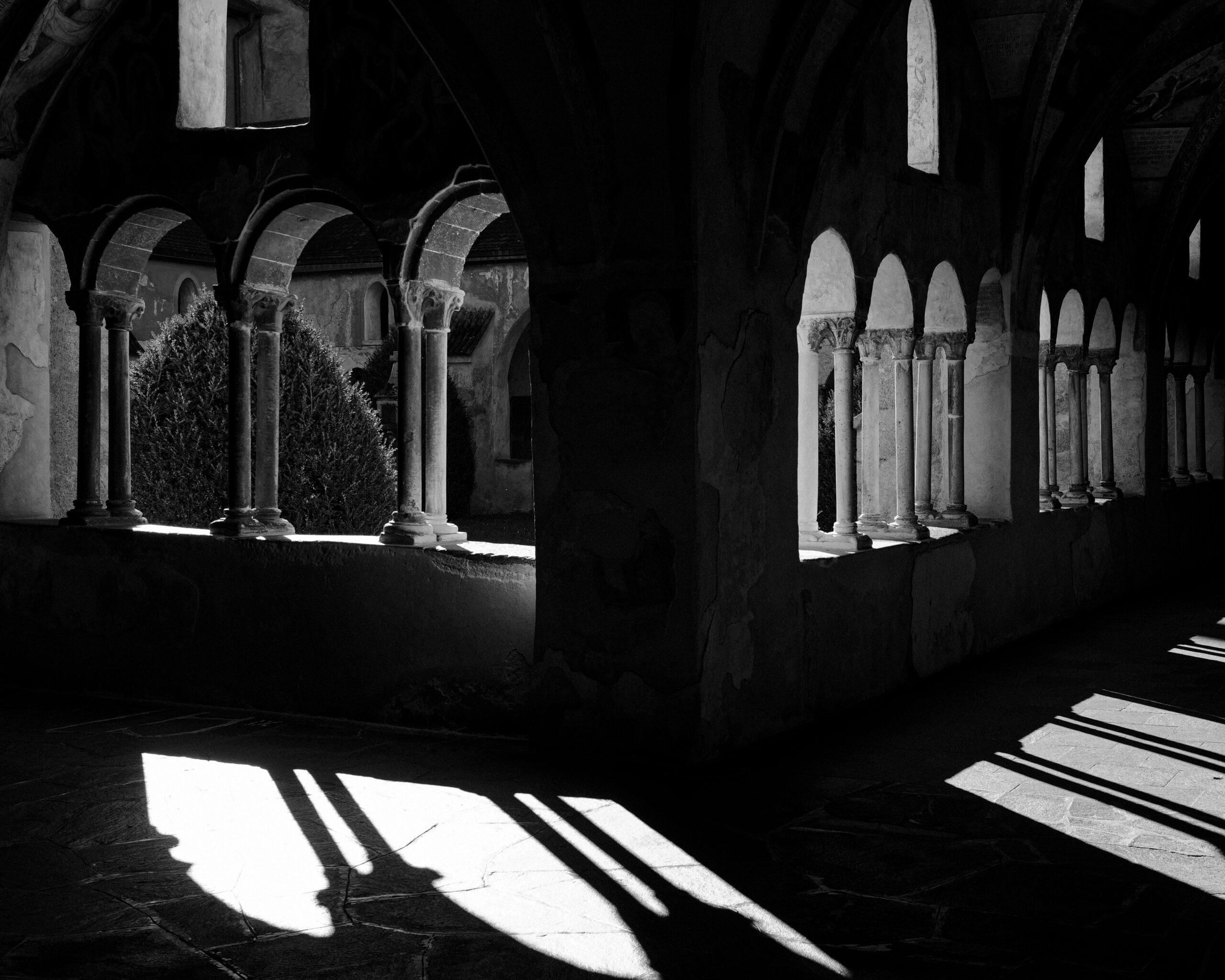 Black and white photo capturing sunlight casting shadows through cloister arches.